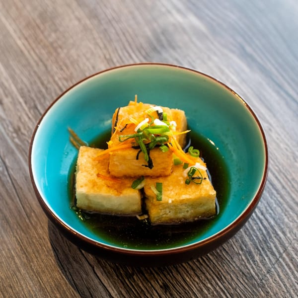 A blue bowl of agedashi tofu, garnished with shredded carrots and green onions, on a wooden table.
