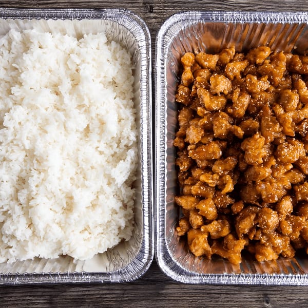 Two foil trays, one with white rice and the other with sesame chicken, on a wooden table.